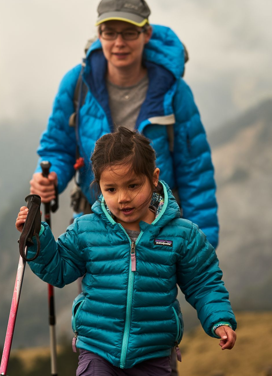 A young girl in a teal puffy jacket walks on a trail holding a pink hiking pole, with an adult in a blue jacket visible behind her in a misty mountain landscape.