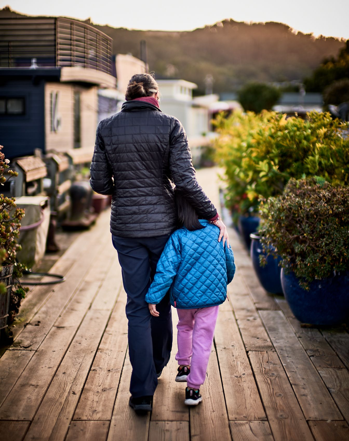 Parent Jen Lumanlan in a dark quilted jacket walks arm-in-arm with daughter Carys wearing a bright blue quilted jacket and pink pants along a wooden dock lined with potted plants