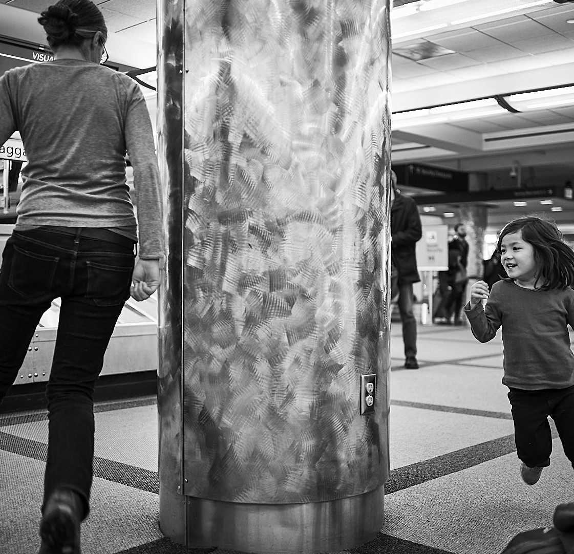 A black and white photo of an adult and child playing hide-and-seek around a large metal column in a train station, with the smiling child peeking out from behind the column.