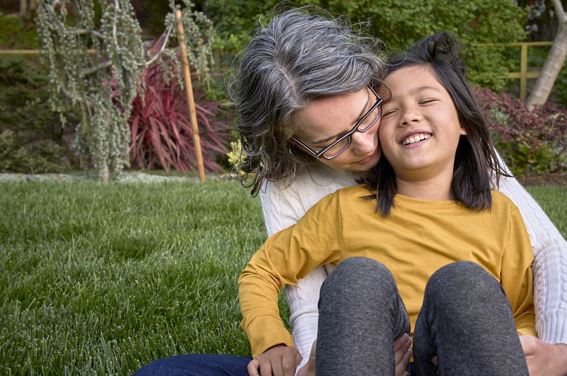 An adult in a yellow shirt and bike helmet stands with a mountain bike in a parking lot with red rock hills in the background, while a child on a smaller bike wearing a yellow-green helmet rides nearby.