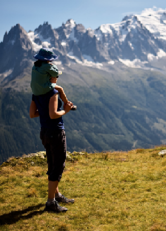 Parent Jen Lumanlan stands on a grassy mountain slope with daughter Carys riding on her shoulders, both looking out at dramatic snow-capped mountain peaks in the distance under a blue sky