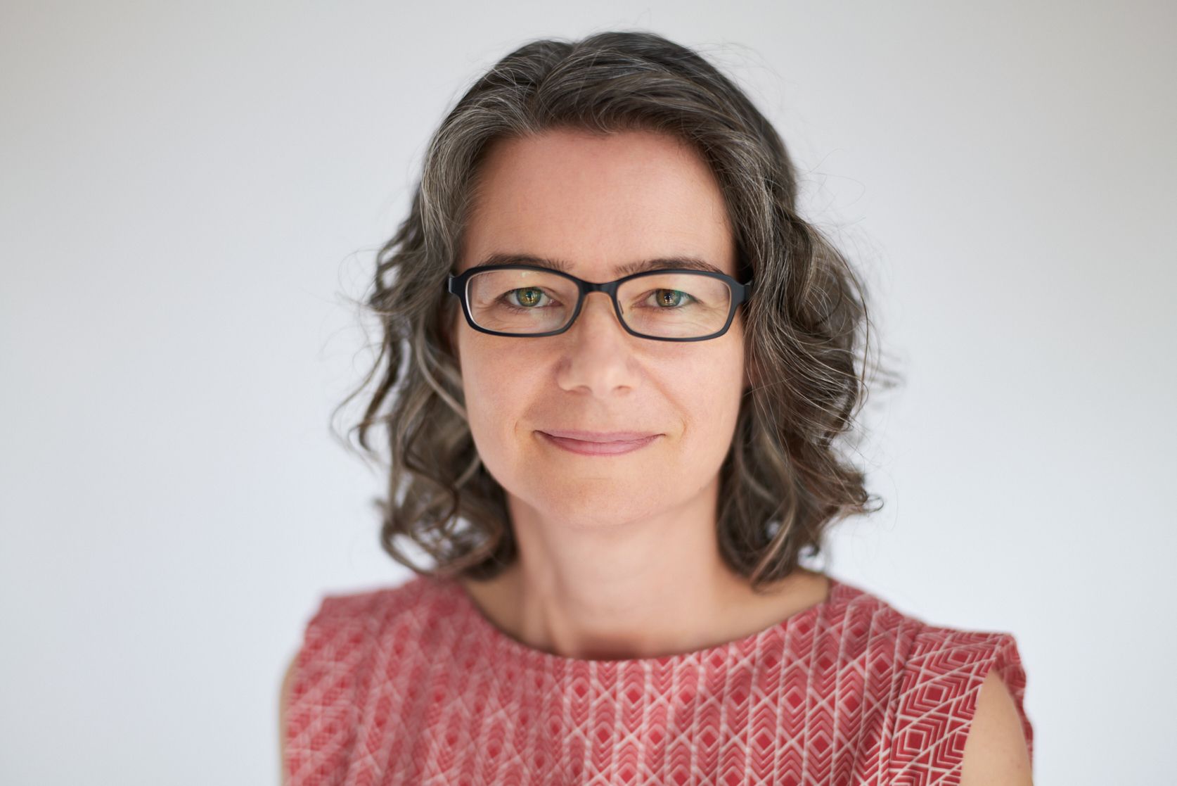 Professional headshot of Jen Lumanlan, a woman with shoulder-length wavy brown hair, black-rimmed glasses, and a warm smile, wearing a red patterned top against a light background