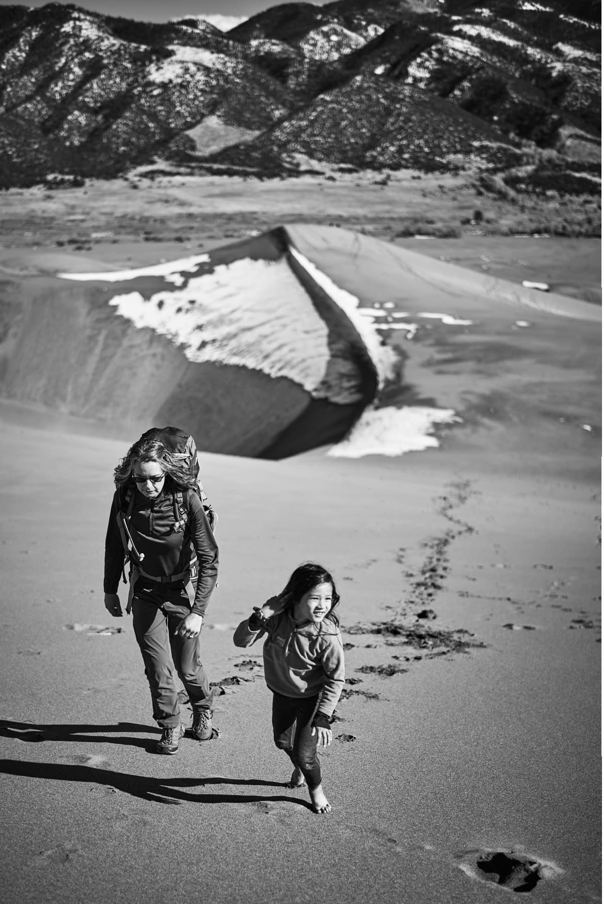 A black and white photograph showing an adult and child hiking up a sand dune. 