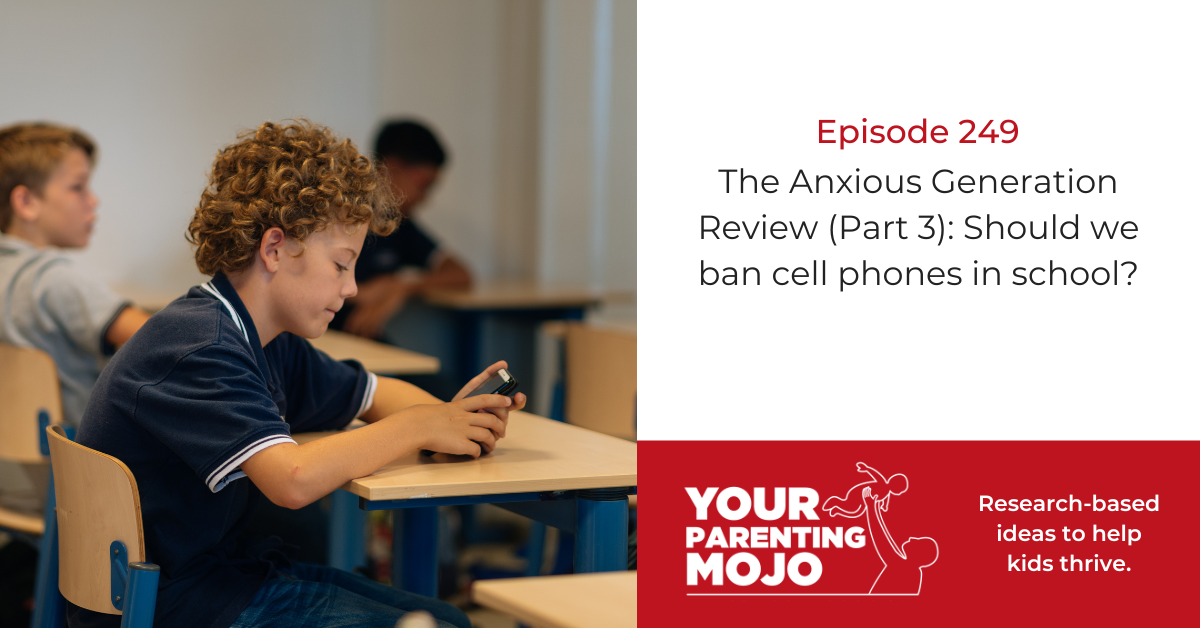A young student with curly hair sits at a school desk, looking down at a cell phone in their hands, with other students visible in the background of the classroom