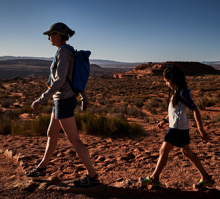 Parent Jen Lumanlan wearing a hat and backpack walks with daughter Carys across red desert terrain at sunset