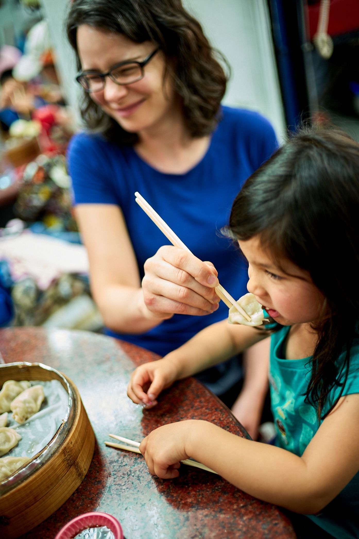 A young girl in a teal puffy jacket walks on a trail holding a pink hiking pole, with an adult in a blue jacket visible behind her in a misty mountain landscape.