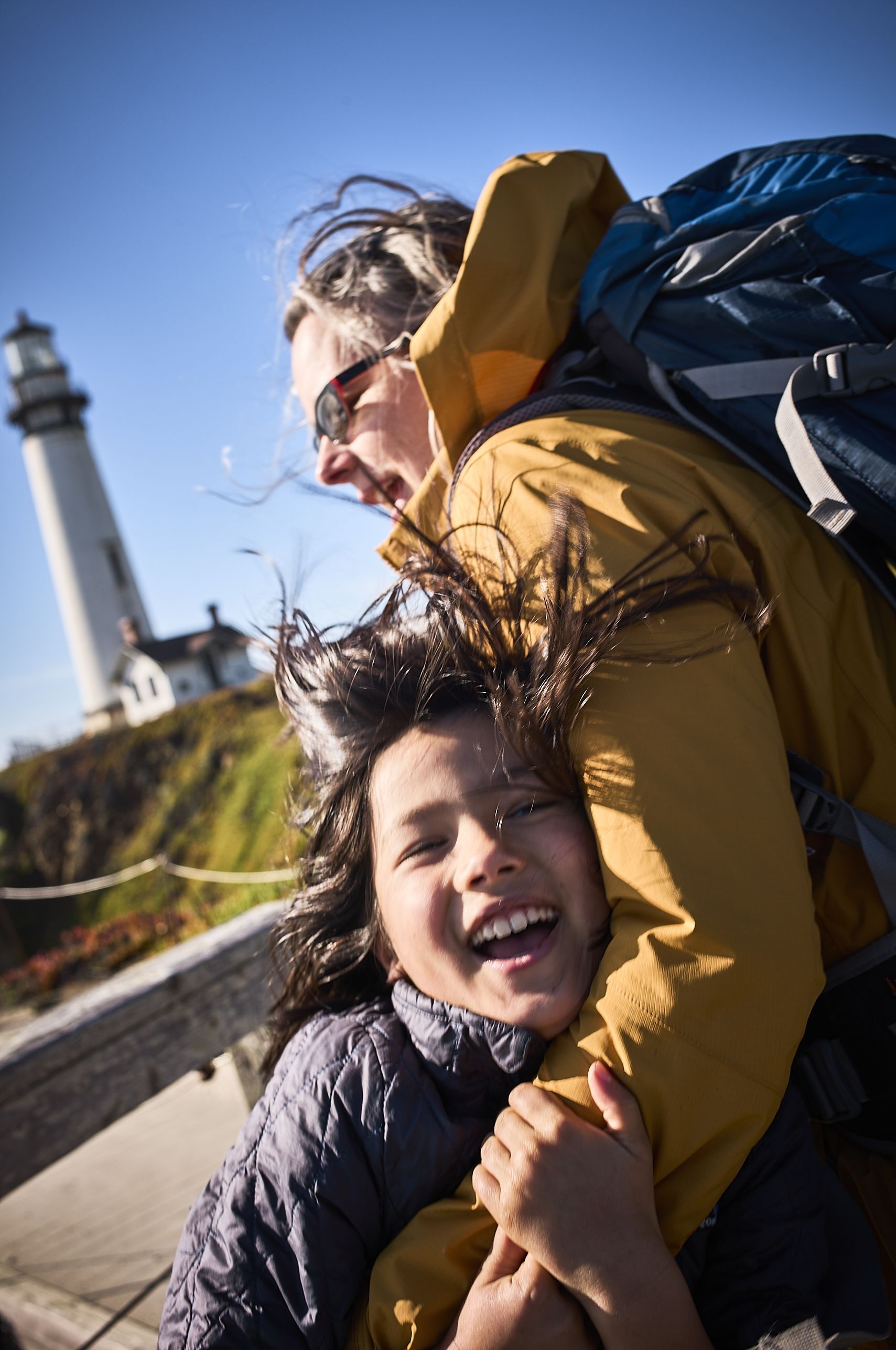 A joyful child in a dark puffy jacket laughs while being held by an adult wearing a yellow jacket and blue backpack, with a white lighthouse visible in the background against a blue sky.