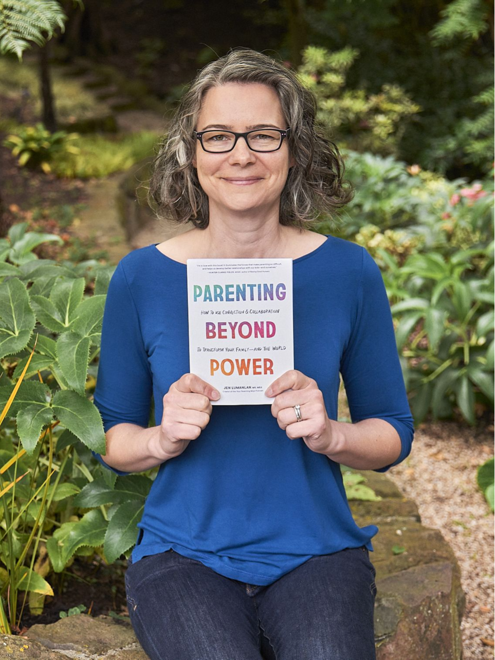 Jen Lumanlan smiles while holding her book "Parenting Beyond Power: How to Use Connection & Collaboration to Transform Your Family—and the World" in a garden setting