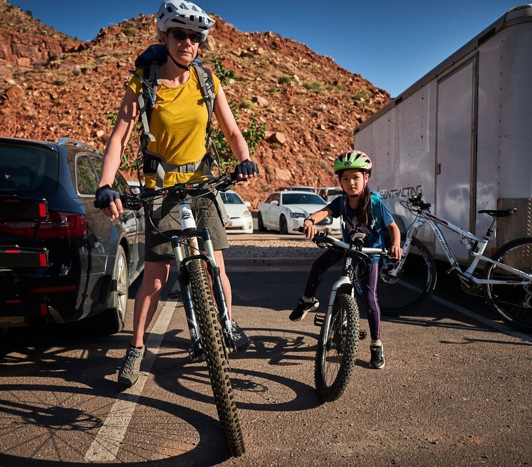 An adult in a yellow shirt and bike helmet stands with a mountain bike in a parking lot with red rock hills in the background, while a child on a smaller bike wearing a yellow-green helmet rides nearby.
