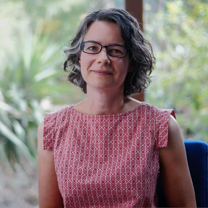 Professional photo of Jen Lumanlan sitting outdoors, wearing black-rimmed glasses and a red geometric patterned sleeveless top, with green foliage softly blurred in the background