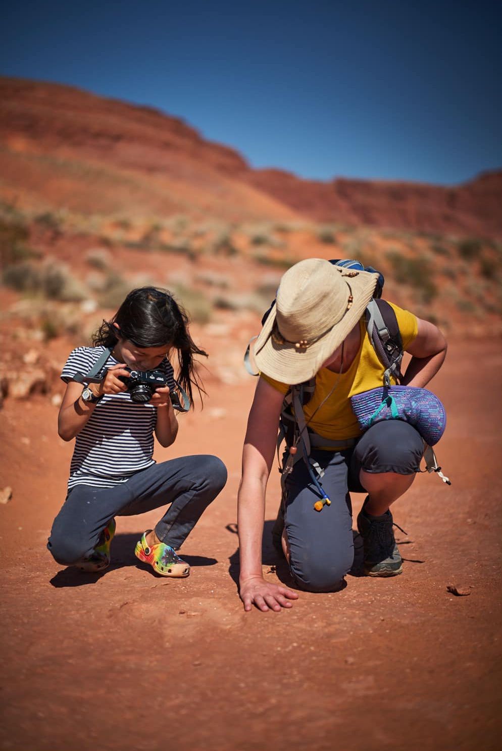A child and an adult are crouched down examining something on the red desert ground.