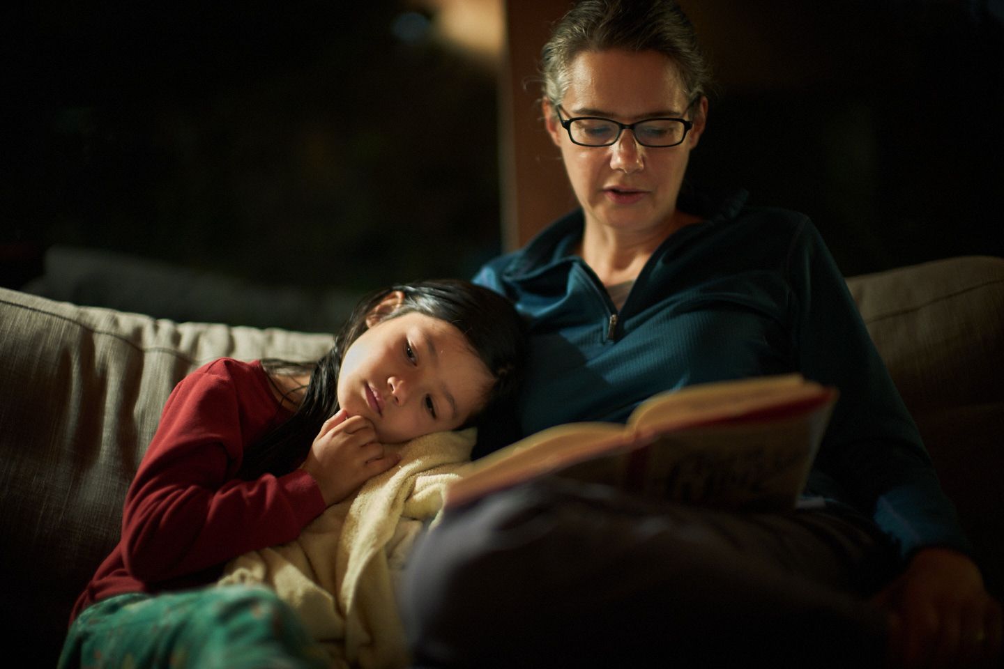en Lumanlan wearing glasses and a teal fleece sits on a couch reading a book while her daughter Carys, wearing a red shirt, rests her head contentedly on Jen's arm