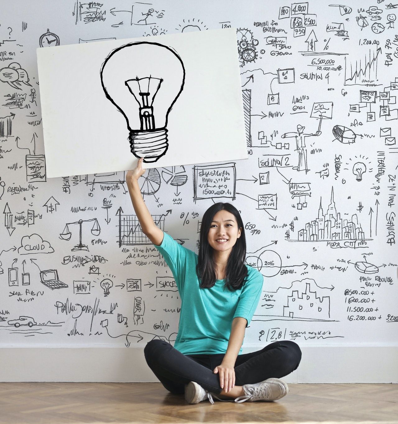 Smiling woman in teal shirt sitting cross-legged holding lightbulb cutout against wall covered in sketched ideas and diagrams representing parenting breakthrough moments