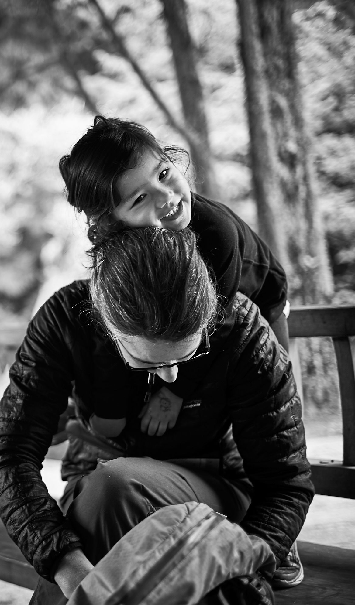 Black and white photo of Jen Lumanlan sitting on wooden steps outdoors while her daughter Carys climbs on her back, both smiling and laughing together.