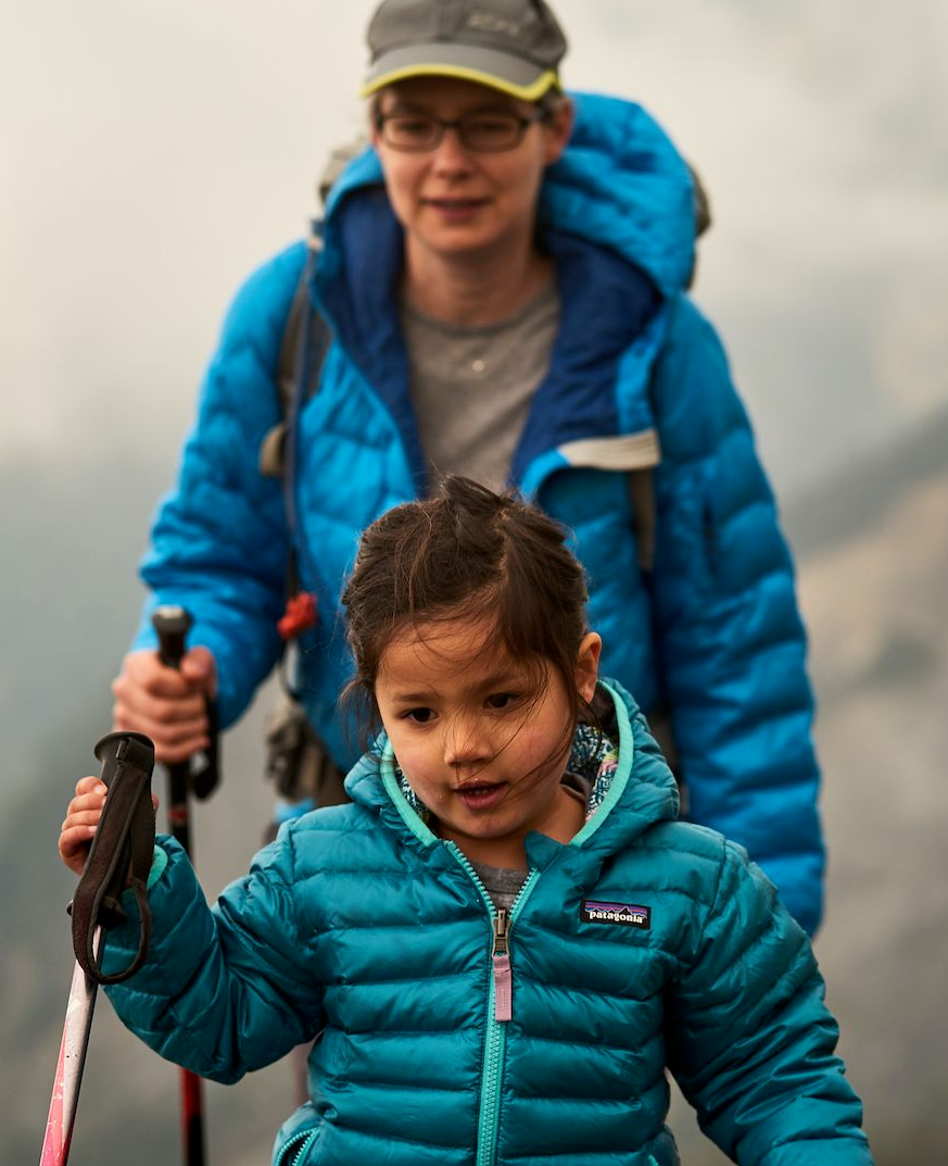 A young girl in a teal puffy jacket walks on a trail holding a pink hiking pole, with an adult in a blue jacket visible behind her in a misty mountain landscape.