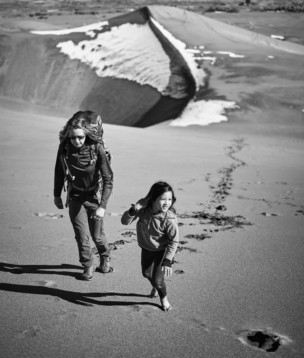 Black and white photo of parent Jen Lumanlan wearing hiking gear and backpack walking alongside barefoot daughter Carys who runs ahead on wet sand