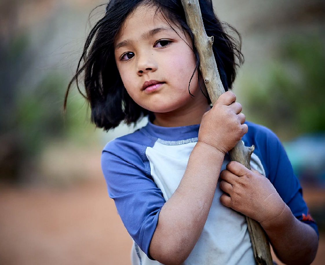 A young girl in a teal puffy jacket walks on a trail holding a pink hiking pole, with an adult in a blue jacket visible behind her in a misty mountain landscape.