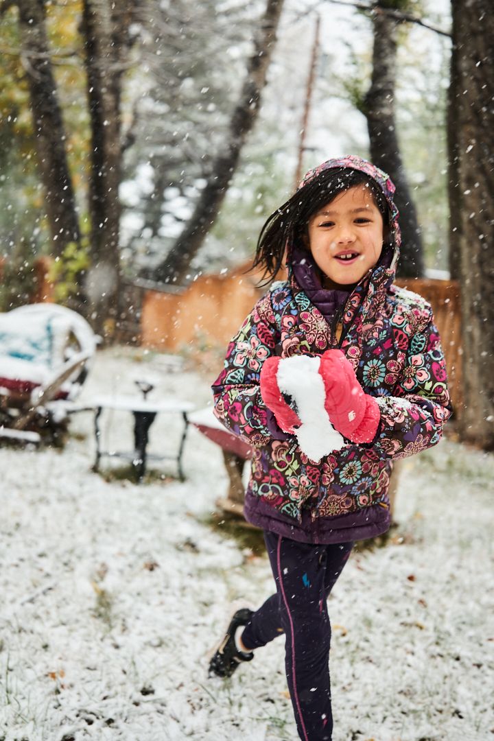Carys, Jen Lumanlan's daughter, running happily through falling snow in a colorful winter coat and red mittens