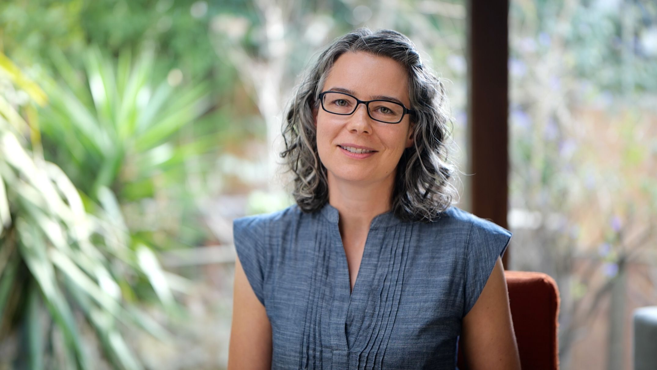 Jen Lumanlan, parenting coach and host of Your Parenting Mojo podcast, smiling while seated in a blue sleeveless top with glasses and shoulder-length wavy hair