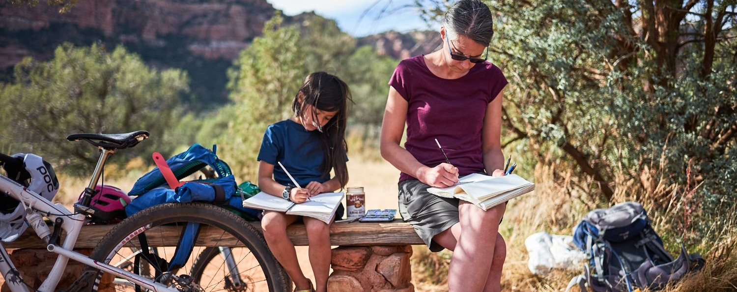 Carys and Jen sit on a wooden bench writing in notebooks, with red rock formations and desert vegetation in the background.