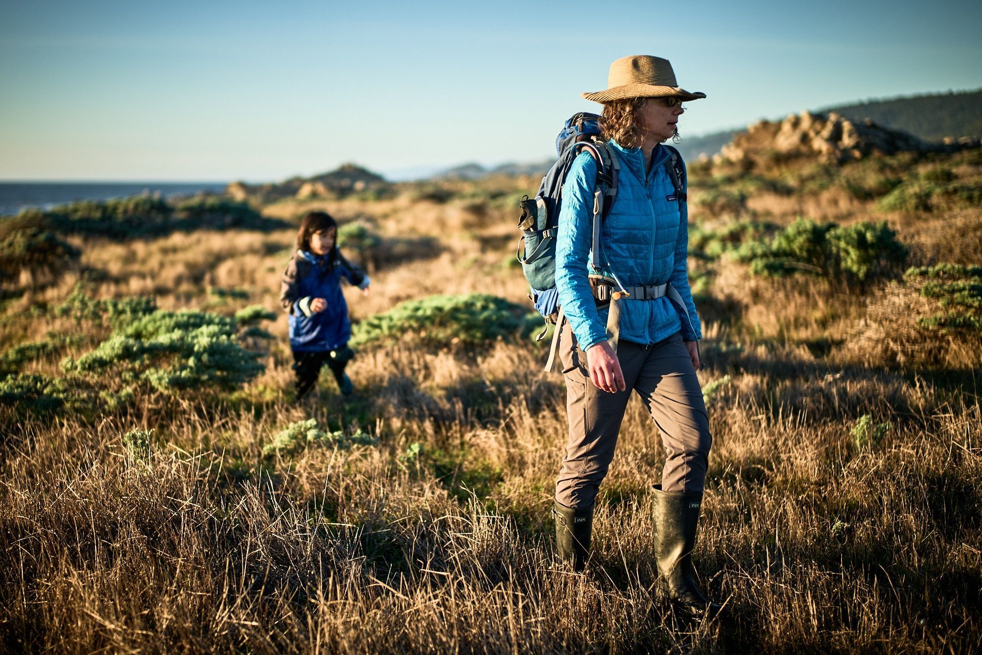 An adult wearing a tan wide-brimmed hat, blue jacket, and backpack walks through coastal grassland with ocean visible in the distance, while a child in a dark blue jacket follows behind.