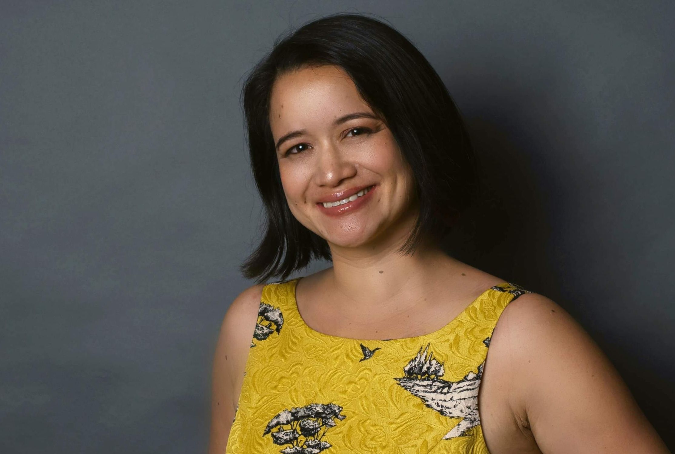 A portrait photograph of a woman wearing a bright yellow sleeveless top with shoulder-length dark hair against a plain gray background.