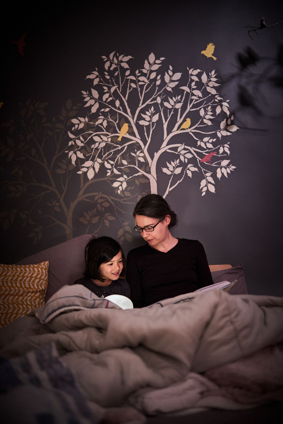 Jen Lumanlan and her daughter Carys reading together in bed under soft lighting with decorative tree wall art, demonstrating connected parenting approach