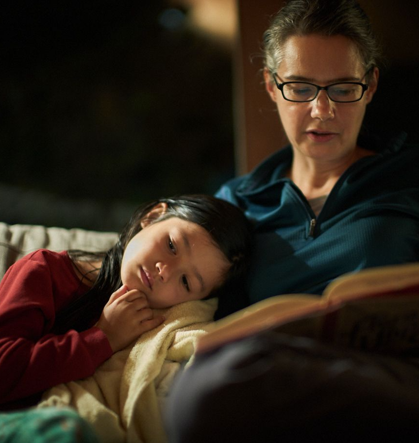 en Lumanlan wearing glasses and a teal fleece sits on a couch reading a book while her daughter Carys, wearing a red shirt, rests her head contentedly on Jen's arm