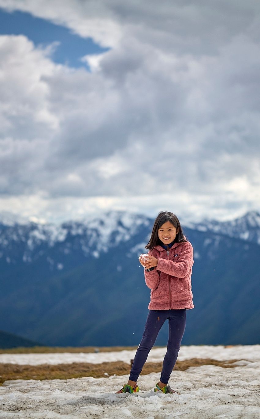 Carys stands smiling on a snowy mountainside wearing a pink fleece jacket, with snow-capped peaks and blue sky behind her.
