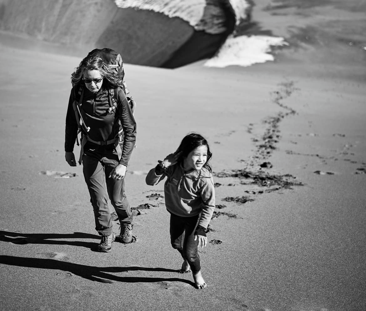 Black and white photo of parent Jen Lumanlan wearing hiking gear and backpack walking alongside barefoot daughter Carys who runs ahead on sand