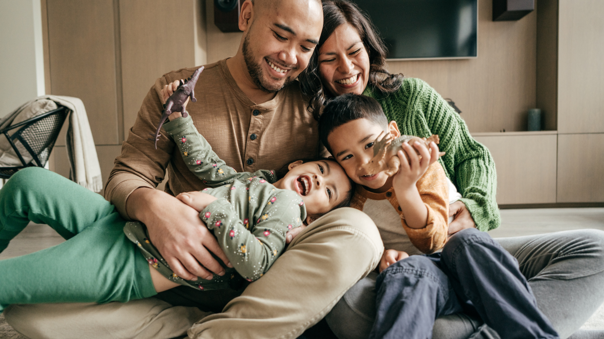 A laughing family of four snuggled together - parents in earth-toned clothing embracing two young children in colorful pajamas, all smiling and appearing relaxed and connected