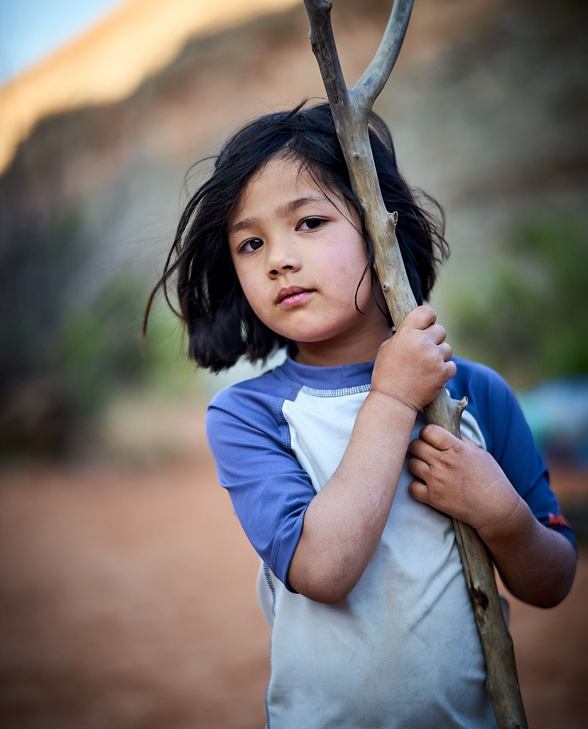 Carys holds a Y-shaped stick over her shoulder, looking directly at the camera with a thoughtful expression