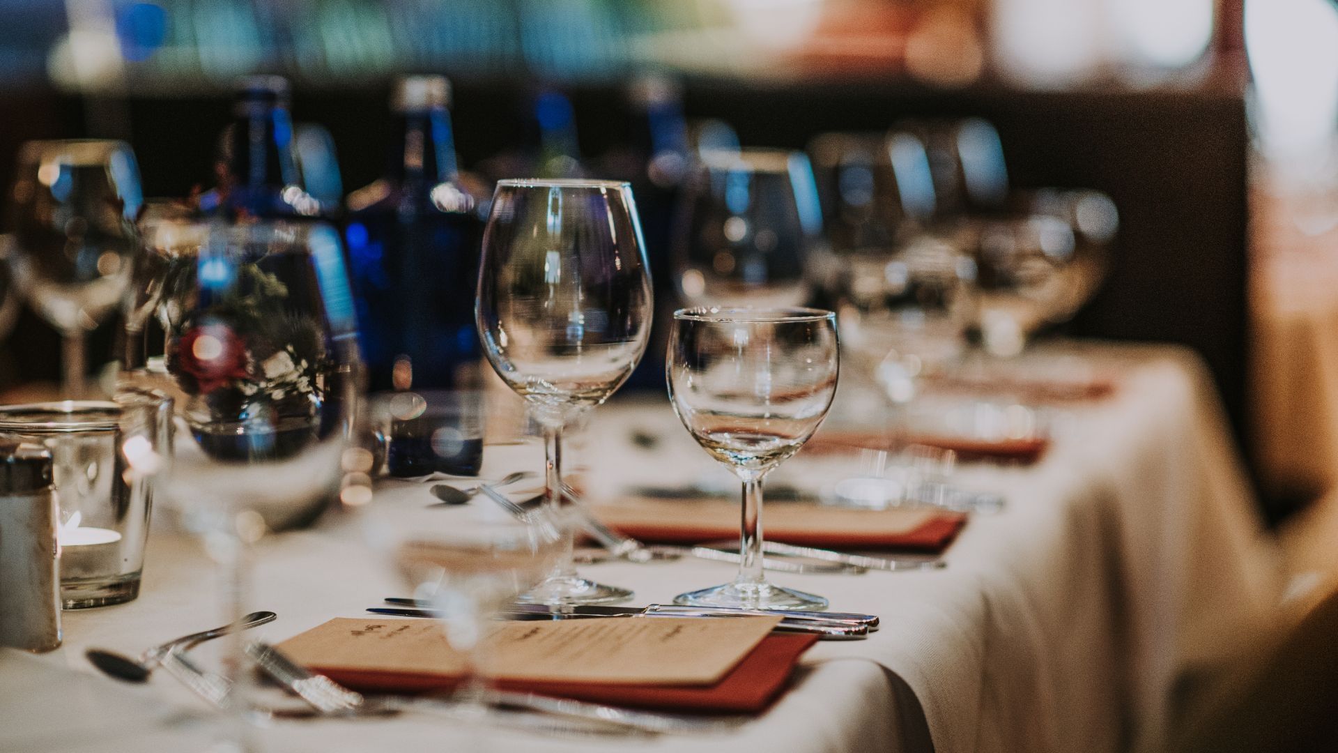 A long table covered in a white tablecloth has been set for dinner with wine glasses, silverware, and paper menus at each seat.