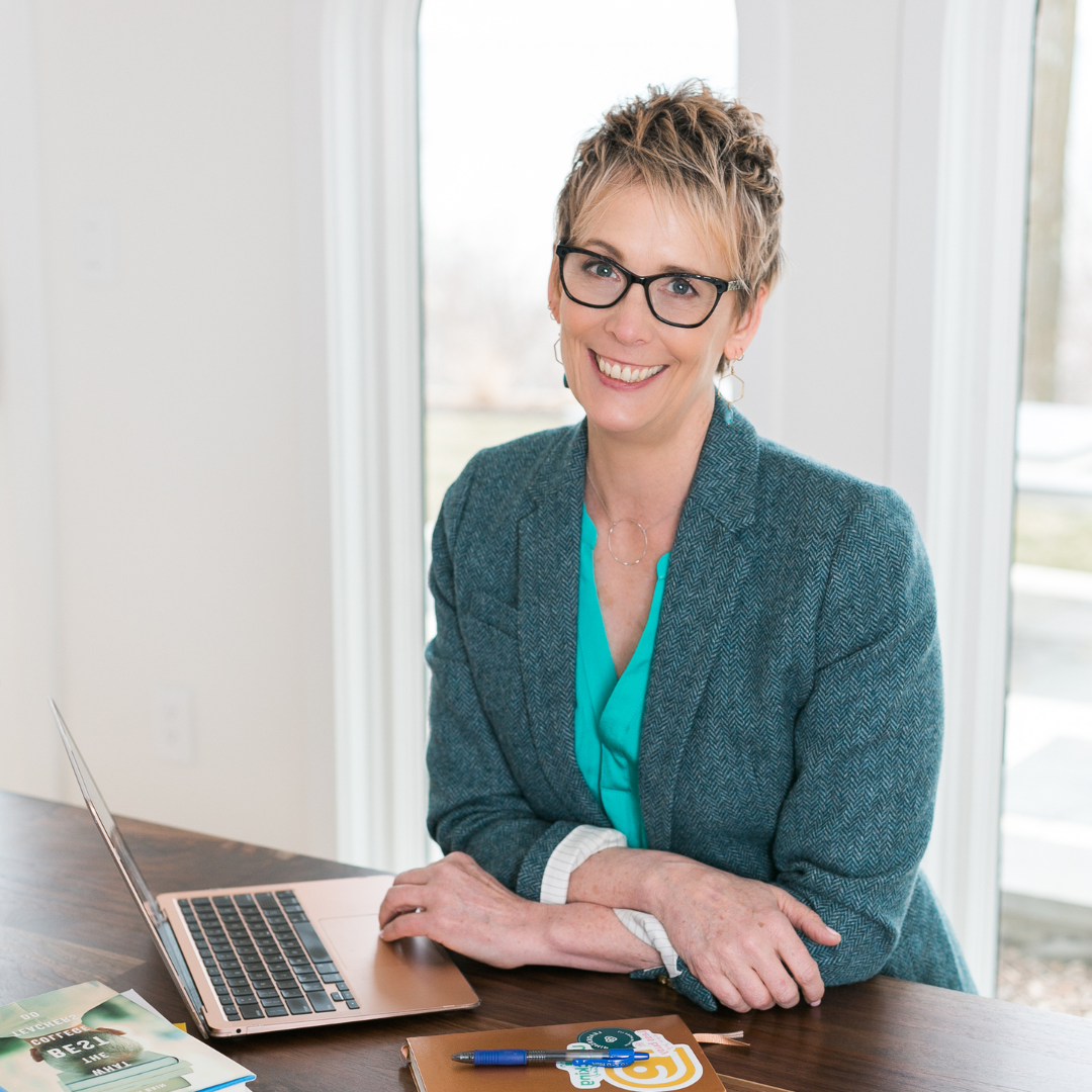 Carole wears a vibrant green top and teal wool blazer with her hands resting on her laptop keyboard. She is smiling at the camera.