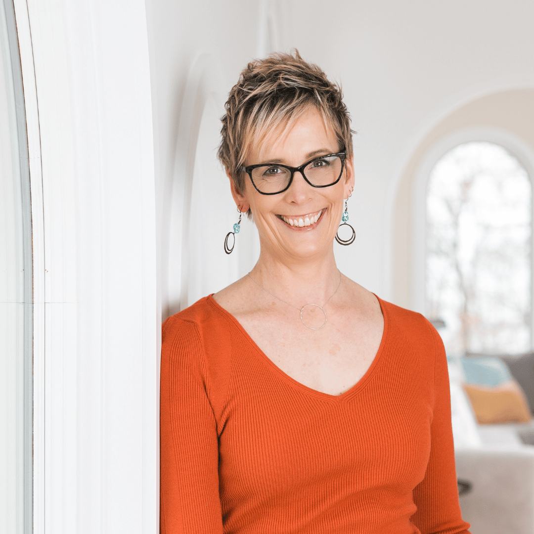 Carole wears a burnt orange v-neck sweater and smiles at the camera.