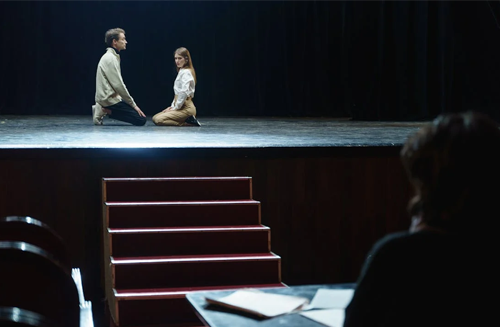 Two individuals seated on a stage, facing an audience.