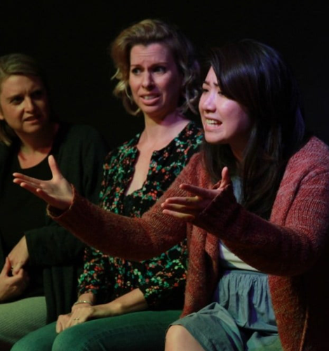 Three women sit on stage, one is gesturing while performing in an acting class